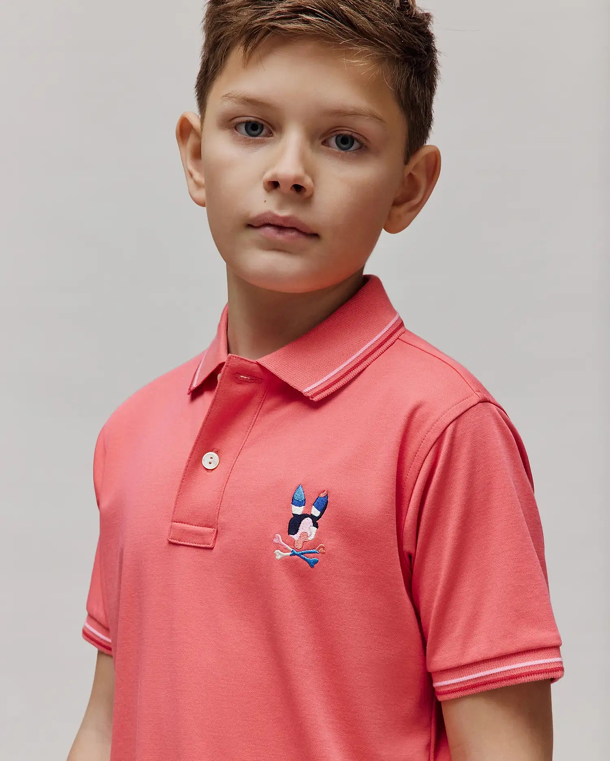 Kids Ara Pique Polo in coral pink, featuring a classic fit, pique knit fabric, ribbed collar and cuffs with white and light pink double stripes, and an embroidered multi-color Psycho Bunny skull logo on the left chest.