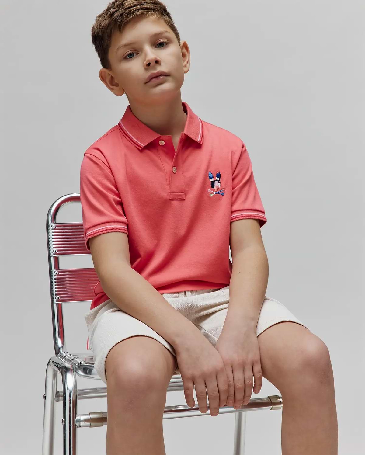 Kids Ara Pique Polo in coral pink, featuring a classic fit, pique texture, and a multi-color embroidered Psycho Bunny skull logo on the left chest. Ribbed collar and cuffs have white and light pink stripes.