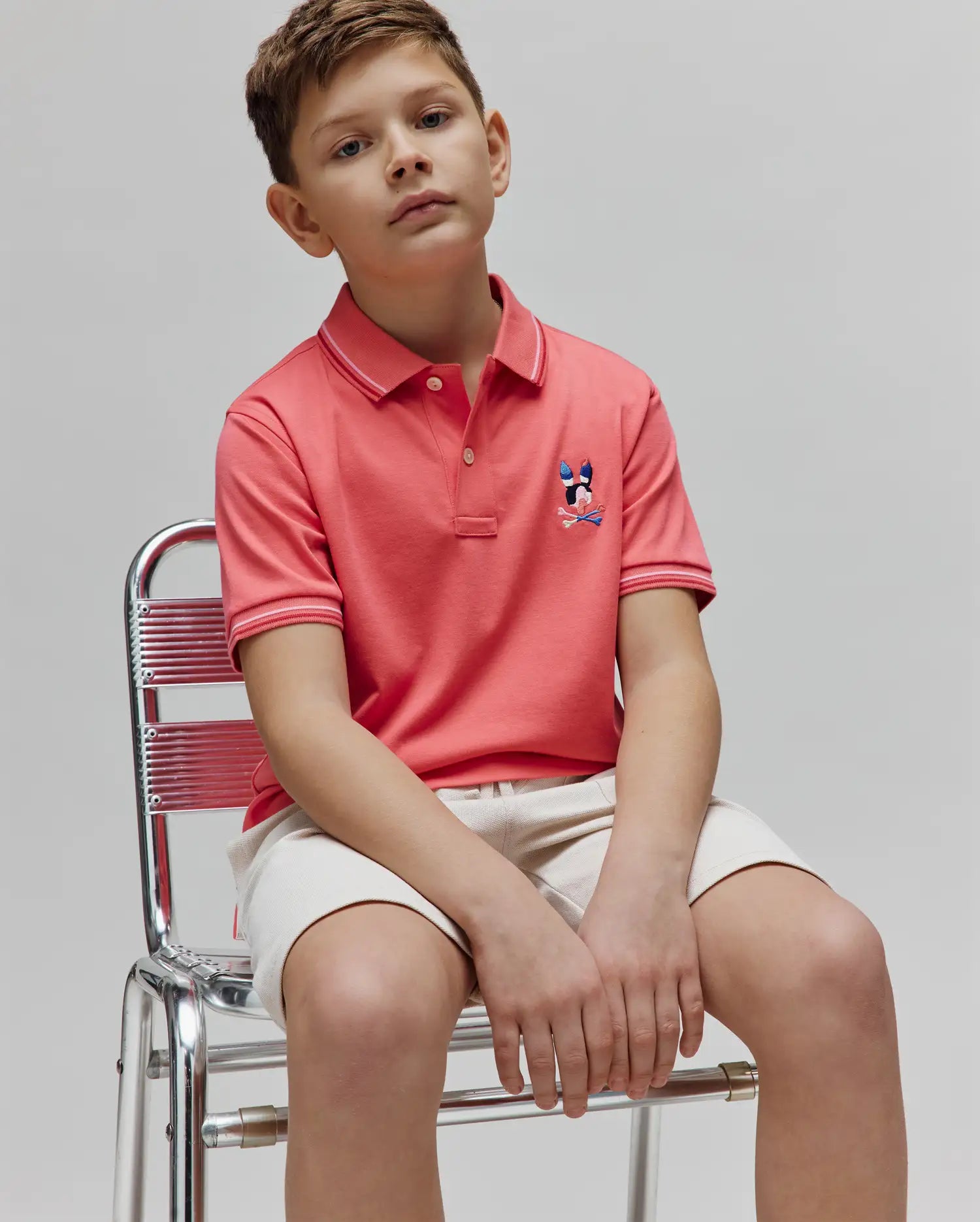 Kids Ara Pique Polo in coral pink, featuring a classic fit, pique texture, and a multi-color embroidered Psycho Bunny skull logo on the left chest. Ribbed collar and cuffs have white and light pink stripes.