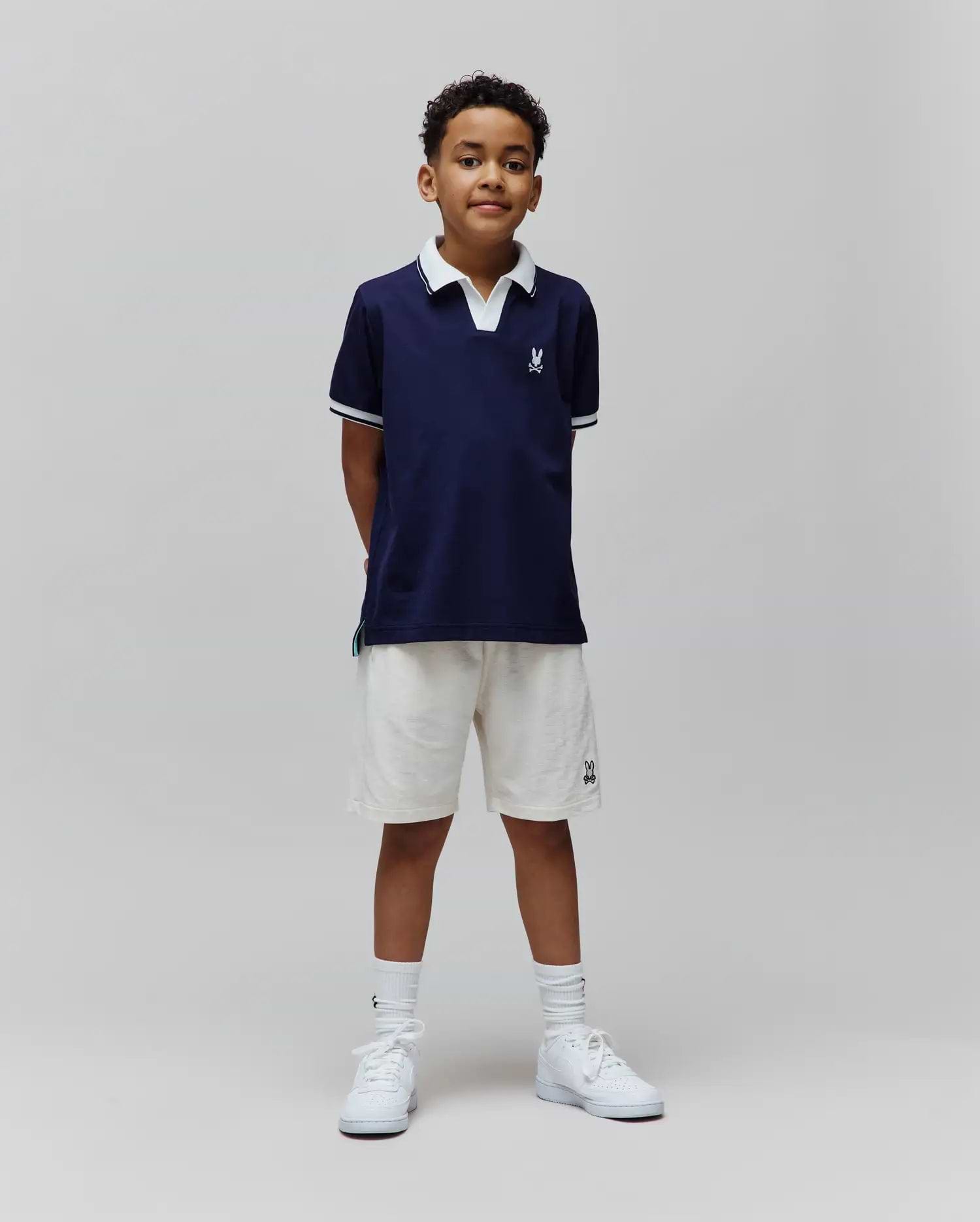 Kids Christiano Pique Polo in navy, classic fit, with white collar, striped cuffs, and embroidered Psycho Bunny skull logo on chest. Cream French terry shorts feature an embroidered bunny logo.