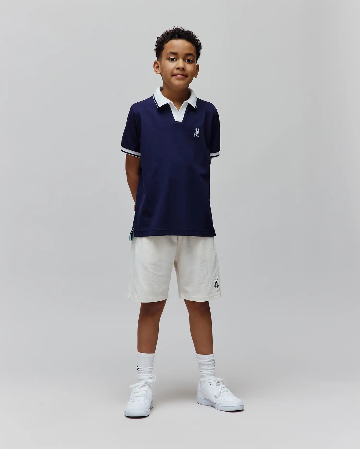 Kids Christiano Pique Polo in navy, classic fit, with white collar, striped cuffs, and embroidered Psycho Bunny skull logo on chest. Cream French terry shorts feature an embroidered bunny logo.