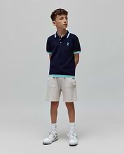 Kids Dax Knit Polo in navy, with light blue and white striped collar, cuffs, and hem. Features a small embroidered light blue Psycho Bunny skull logo on the chest. Paired with light beige knit shorts.