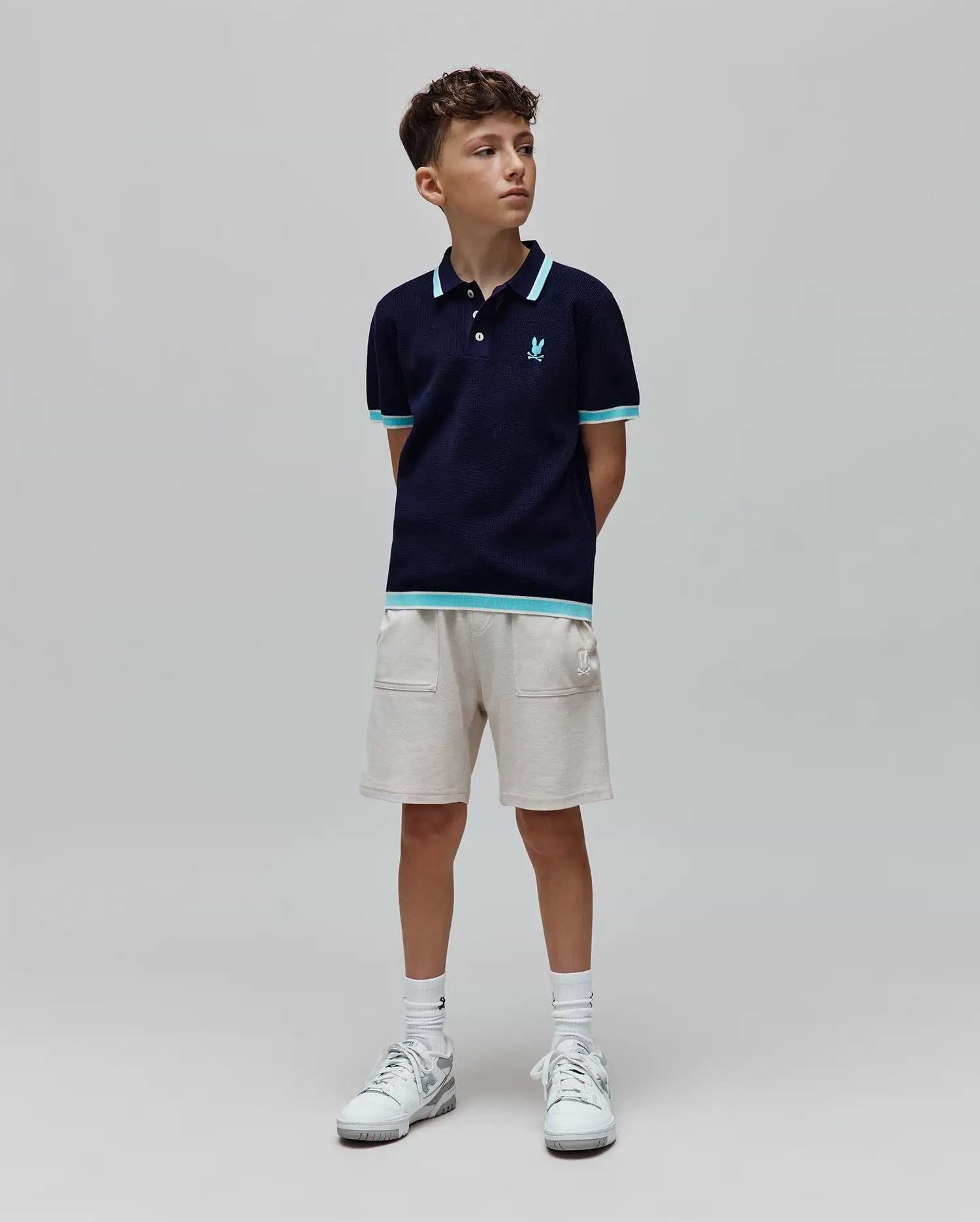 Kids Dax Knit Polo in navy, with light blue and white striped collar, cuffs, and hem. Features a small embroidered light blue Psycho Bunny skull logo on the chest. Paired with light beige knit shorts.
