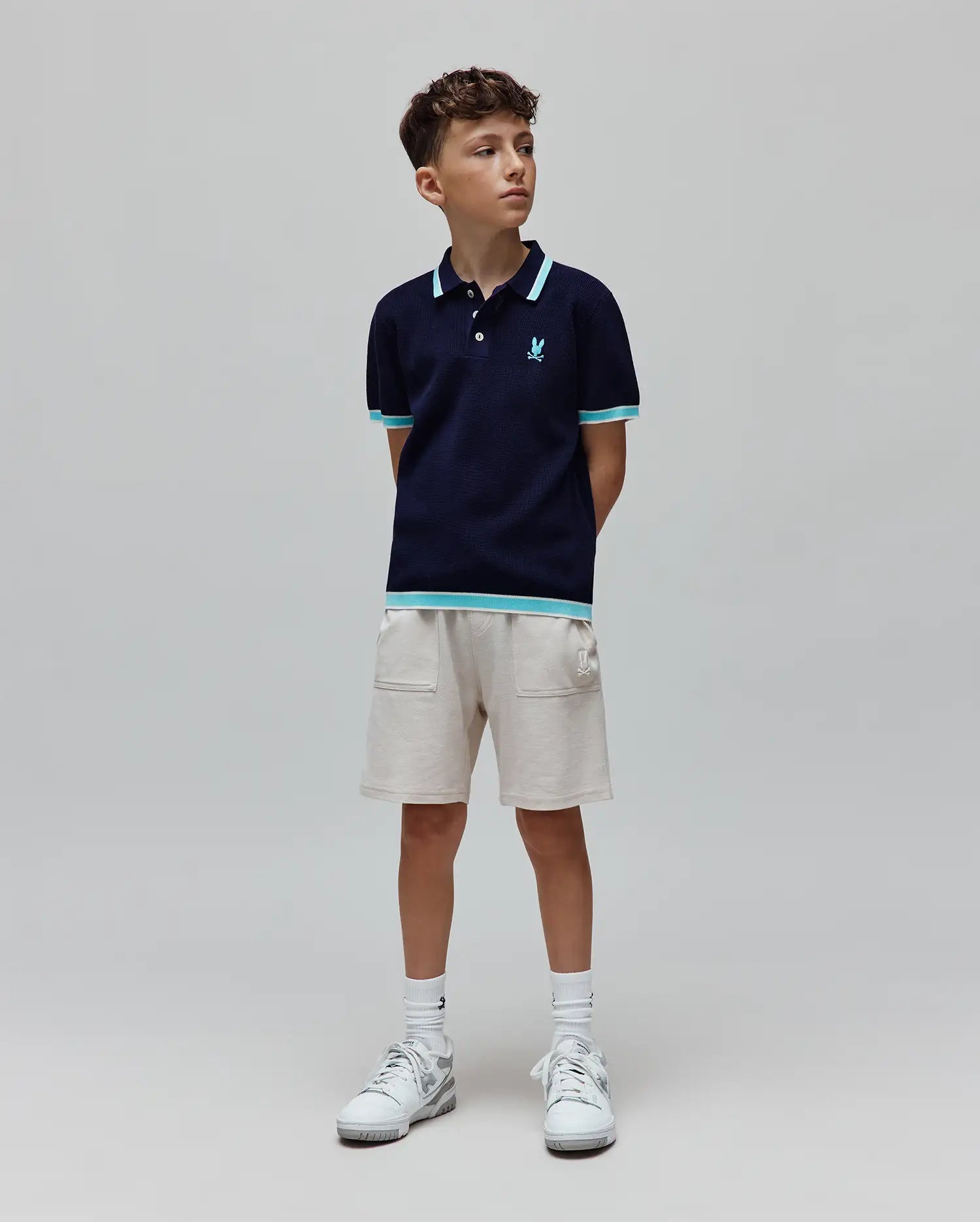Kids Dax Knit Polo in navy, with light blue and white striped collar, cuffs, and hem. Features a small embroidered light blue Psycho Bunny skull logo on the chest. Paired with light beige knit shorts.