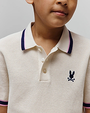 Kids Dax Knit Polo in cream, a classic fit textured knit polo with a two-button placket, small navy embroidered Psycho Bunny skull logo on the left chest, and ribbed collar/cuffs with navy and pink stripes.