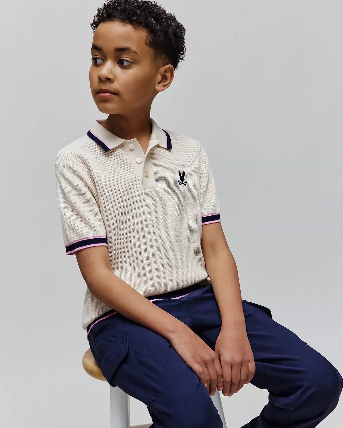 Kids Dax Knit Polo in cream waffle knit with navy and pink striped collar and cuffs, featuring a small embroidered Psycho Bunny skull logo on the left chest.