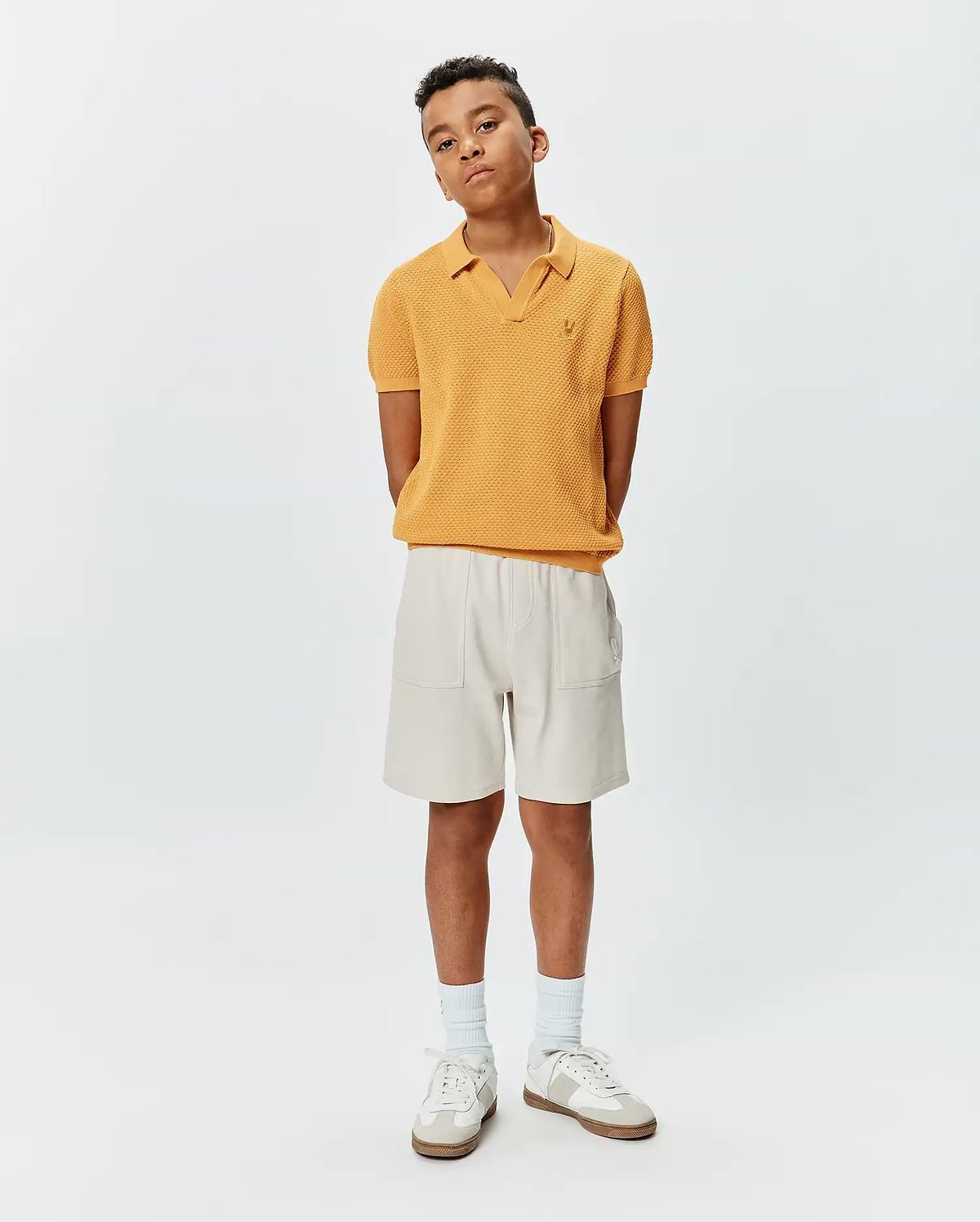 Kids Devon Knit Polo in mustard yellow, featuring a textured knit fabric, V-neck, and small embroidered Psycho Bunny skull logo on the left chest, paired with relaxed fit light beige shorts with patch pockets.