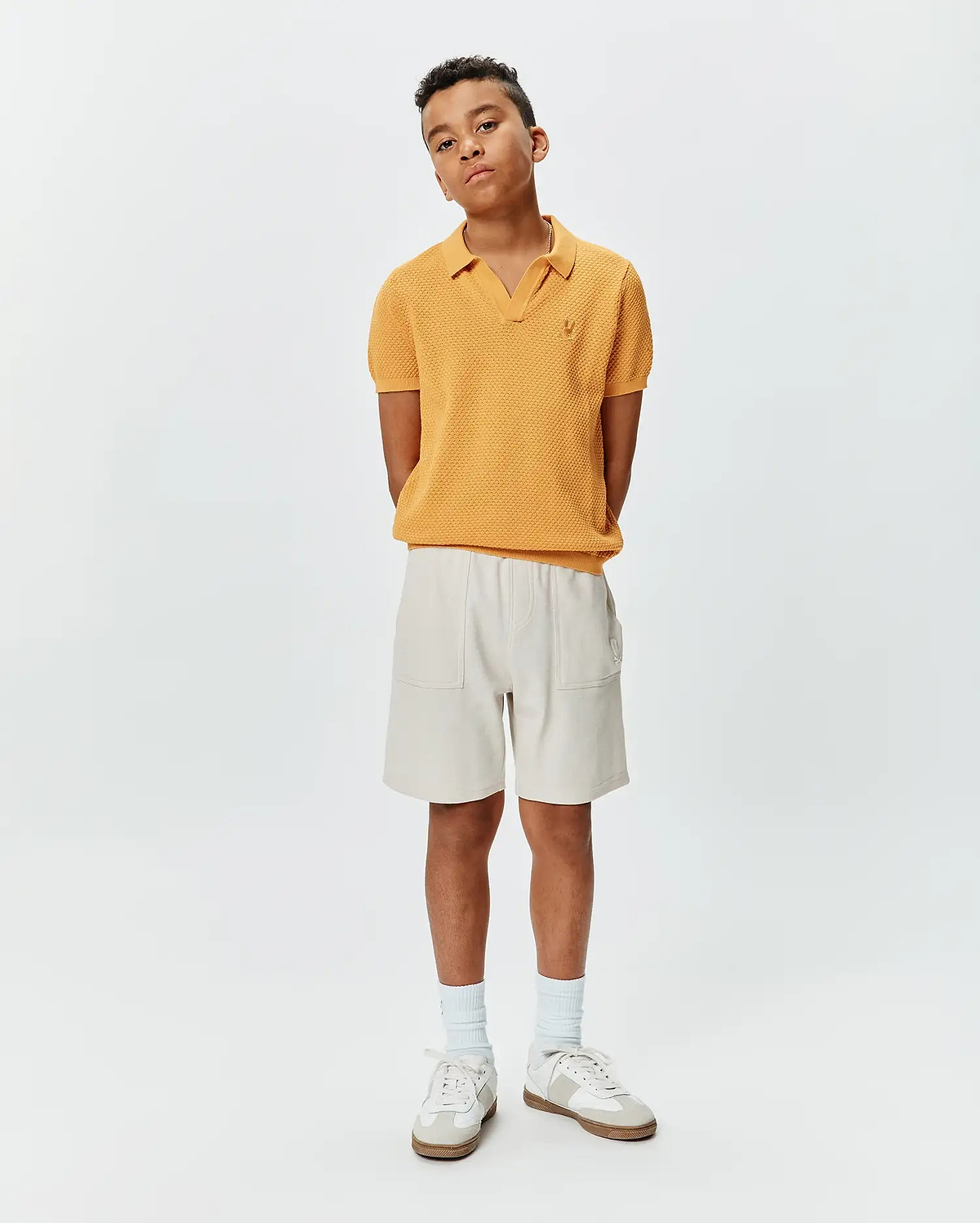 Kids Devon Knit Polo in mustard yellow, featuring a textured knit fabric, V-neck, and small embroidered Psycho Bunny skull logo on the left chest, paired with relaxed fit light beige shorts with patch pockets.
