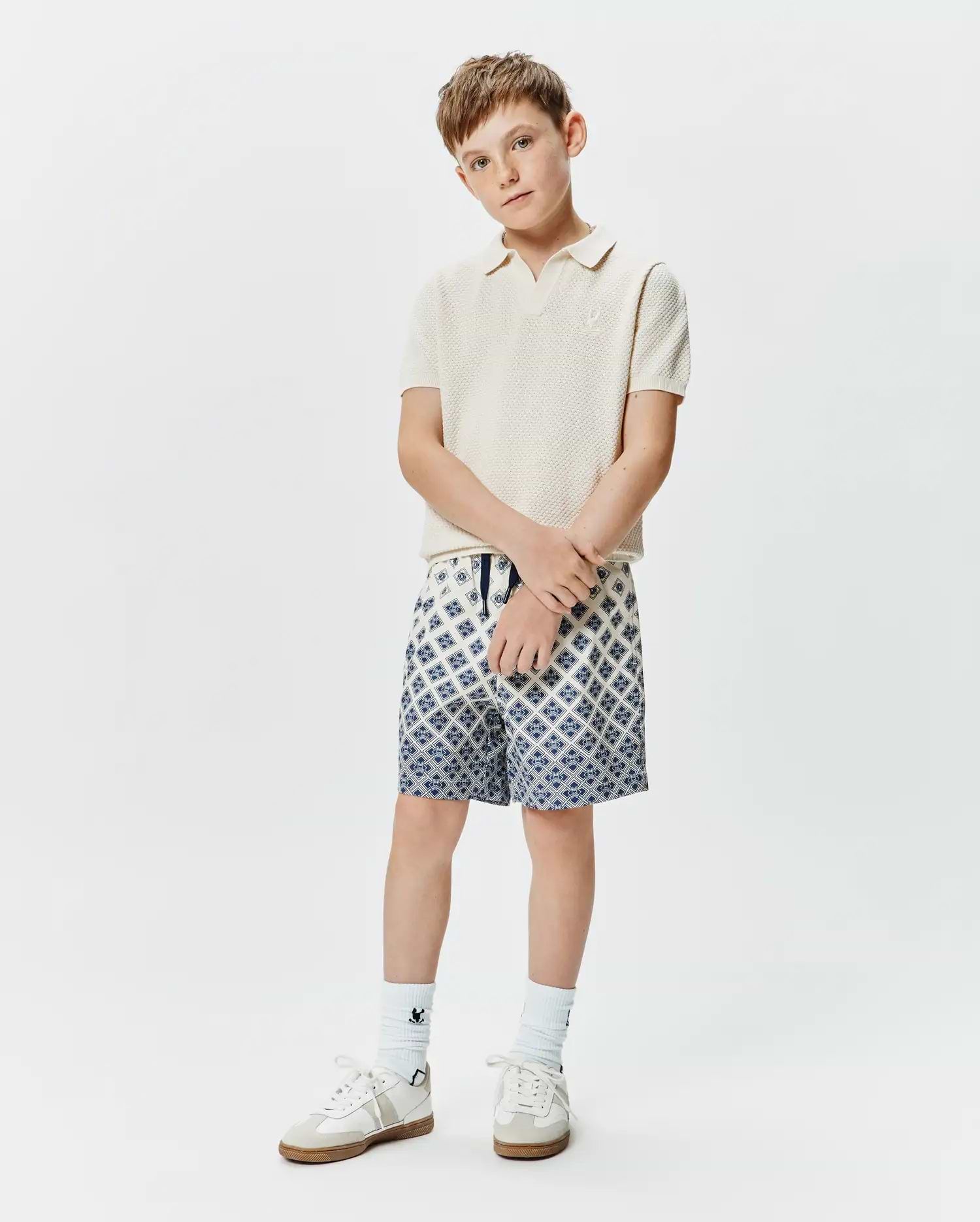 Kids Devon Knit Polo in cream, featuring a textured knit fabric and small embroidered Psycho Bunny logo on the chest, paired with blue and white geometric pattern shorts.