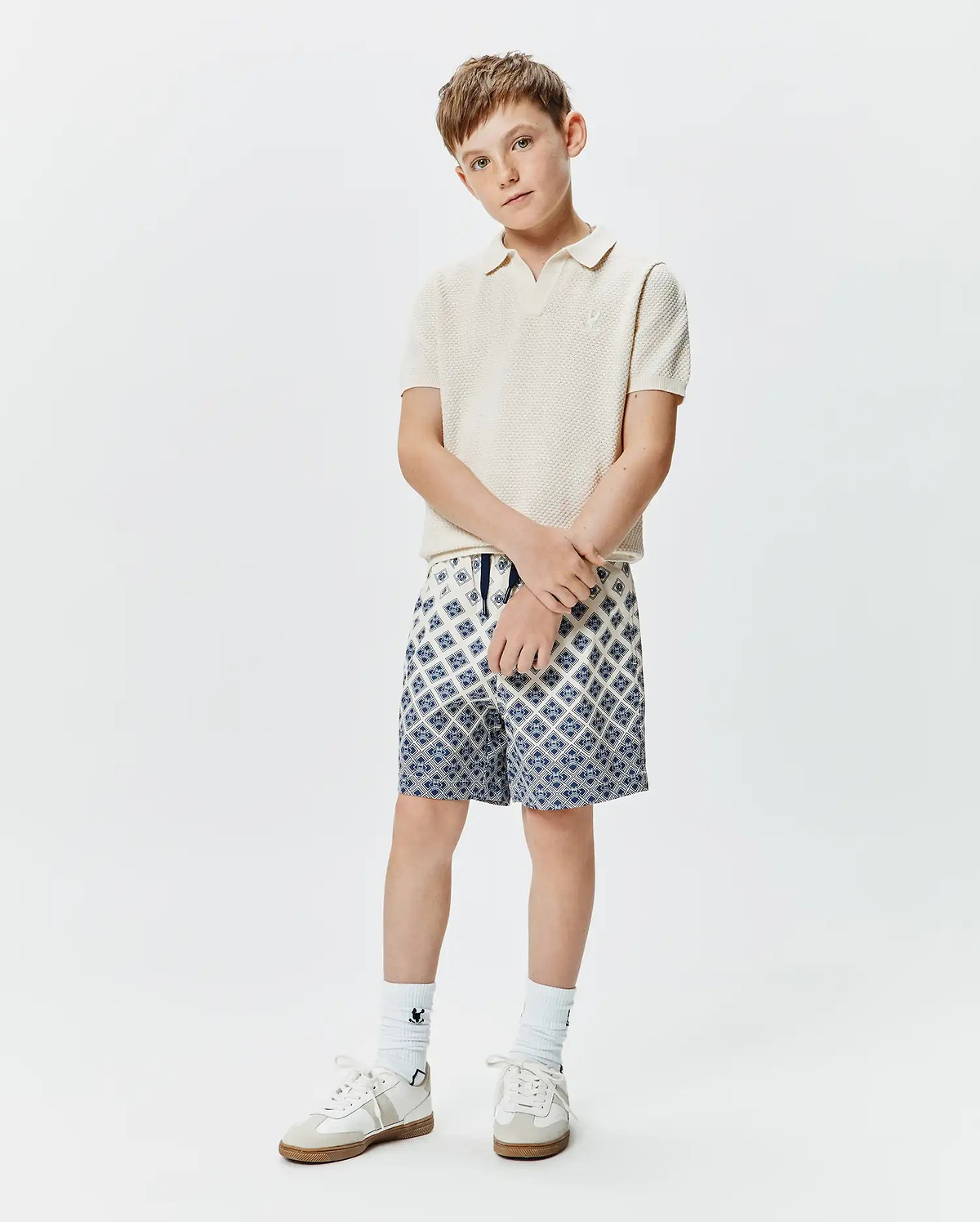 Kids Devon Knit Polo in cream, featuring a textured knit fabric and small embroidered Psycho Bunny logo on the chest, paired with blue and white geometric pattern shorts.