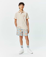 Beige KIDS HOLDEN PIQUE POLO with orange trim and embroidered Psycho Bunny logo on left chest, paired with blue and orange geometric patterned shorts featuring integrated bunny skull motifs.