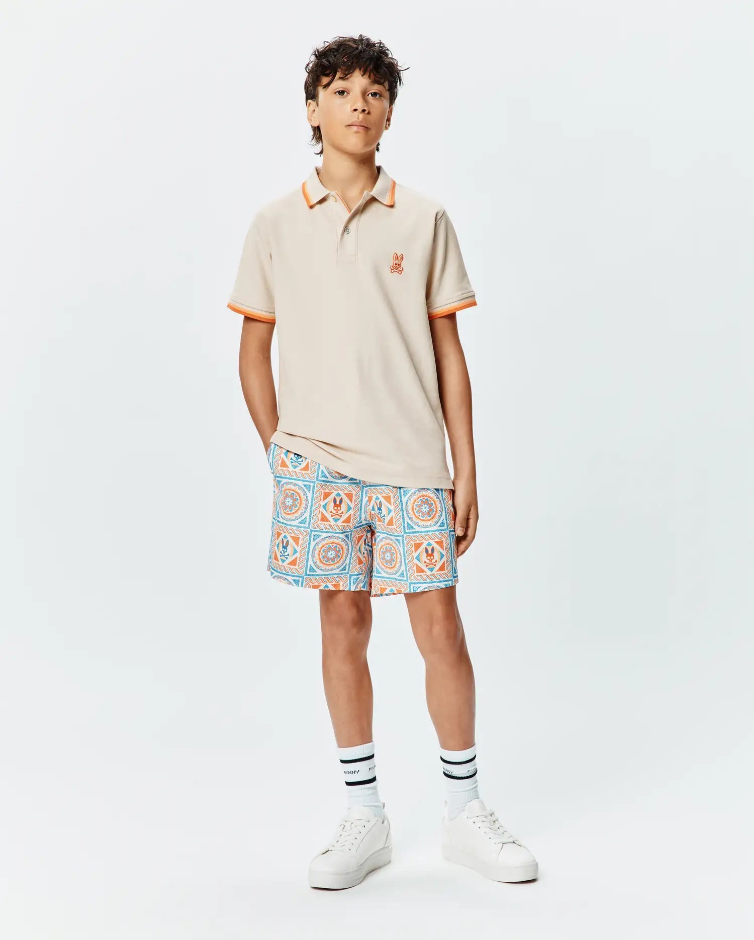 Beige KIDS HOLDEN PIQUE POLO with orange trim and embroidered Psycho Bunny logo on left chest, paired with blue and orange geometric patterned shorts featuring integrated bunny skull motifs.