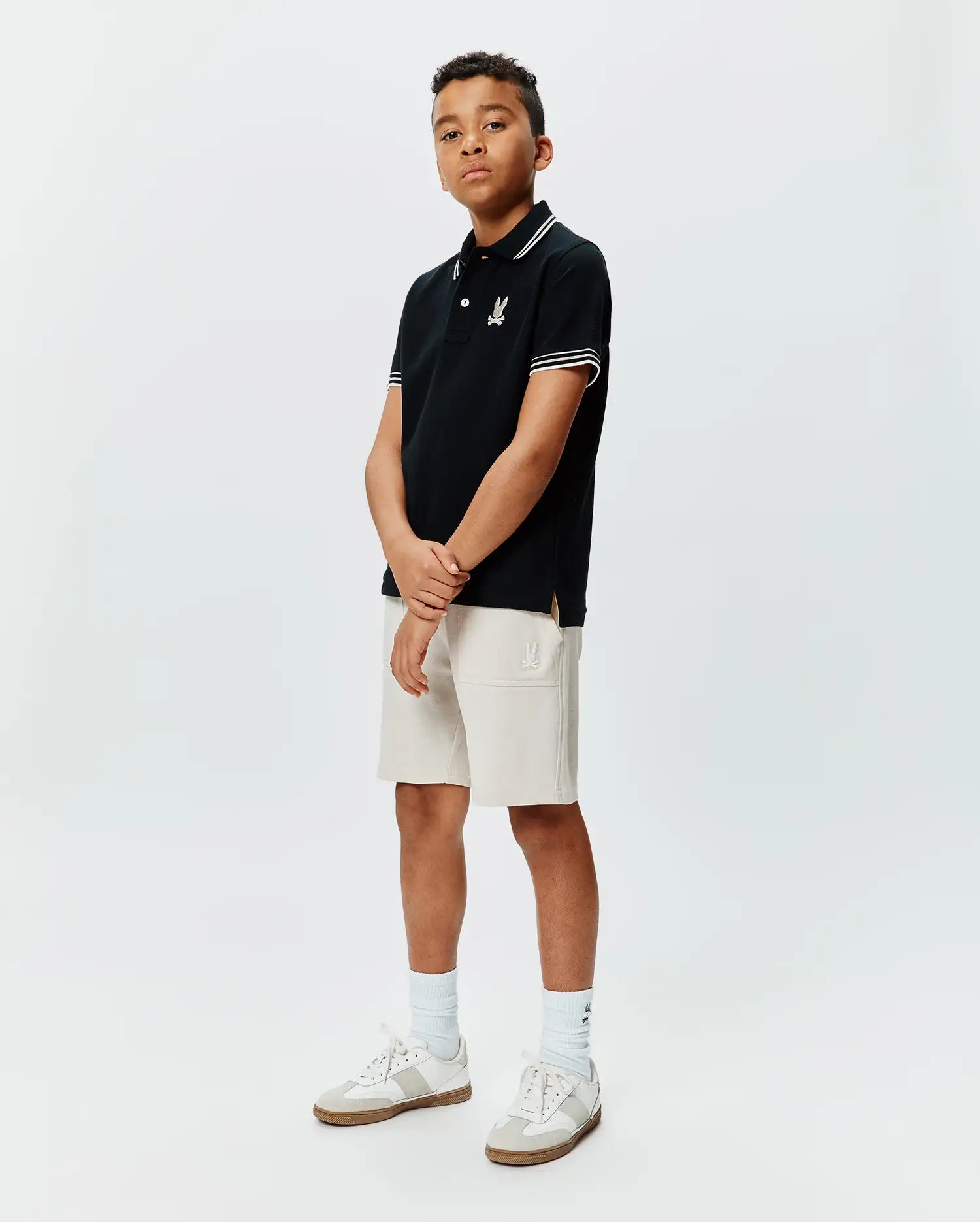 Kids Brad Pique Polo in black with white collar and sleeve tipping, featuring an embroidered Psycho Bunny skull logo on the left chest, paired with light beige knit shorts with an embroidered Psycho Bunny logo.