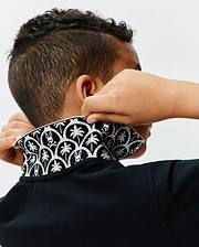 Kids Brad Pique Polo in classic black, featuring a textured pique fabric and a bold, contrasting white pattern on the collar with repeating palm trees and iconic Psycho Bunny skull logos.