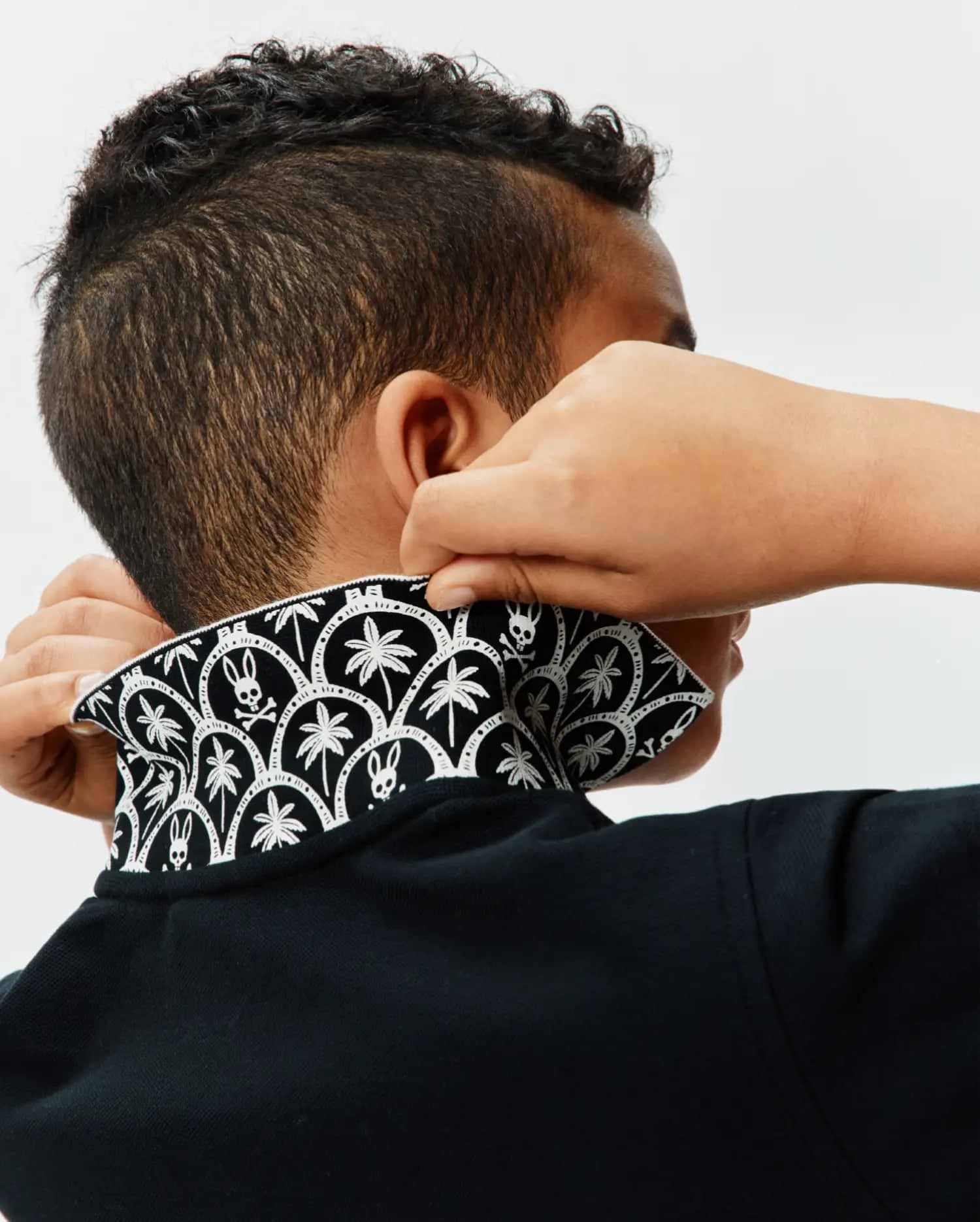 Kids Brad Pique Polo in classic black, featuring a textured pique fabric and a bold, contrasting white pattern on the collar with repeating palm trees and iconic Psycho Bunny skull logos.