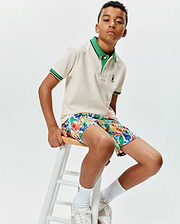 Kids Sunny Pique Polo in cream pique with a green collar and striped cuffs, featuring a small embroidered Psycho Bunny logo, paired with multi-color abstract print shorts. (3)