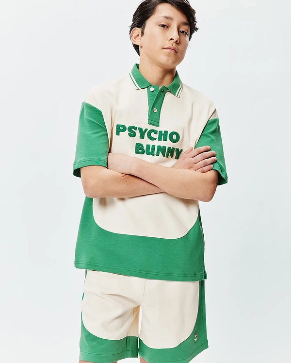 Kids Kelly Oversized Polo in cream and green color block. Features a striped collar, two-button placket, and large textured PSYCHO BUNNY chest embroidery. Matching shorts have a small bunny skull logo.