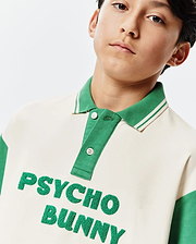 Kids Kelly Oversized Polo, a cream and green color-block jersey polo featuring a ribbed striped collar, two-button placket, and large textured green embroidered Psycho Bunny text on the chest.