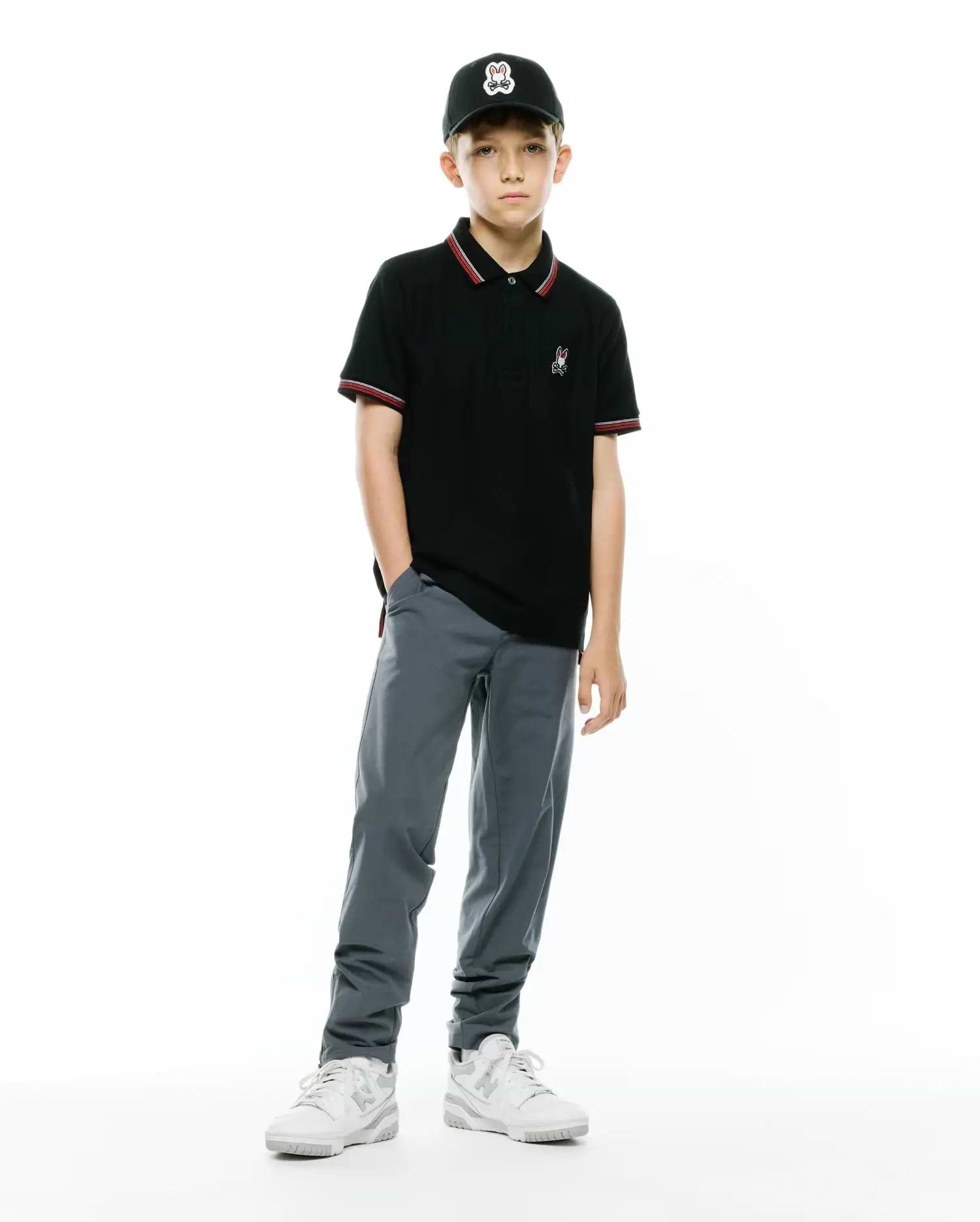 KIDS JAYDON PIQUE POLO, a black classic fit pique polo with red/white striped collar/cuffs, featuring a small embroidered Psycho Bunny logo. Worn with grey pants, white sneakers, and a black cap with a large Psycho Bunny logo.