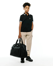 Kids Timmy Pique Polo in classic black pique with white striped collar and cuffs, featuring a small embroidered white Psycho Bunny skull logo on the left chest, styled with khaki pants. (4)