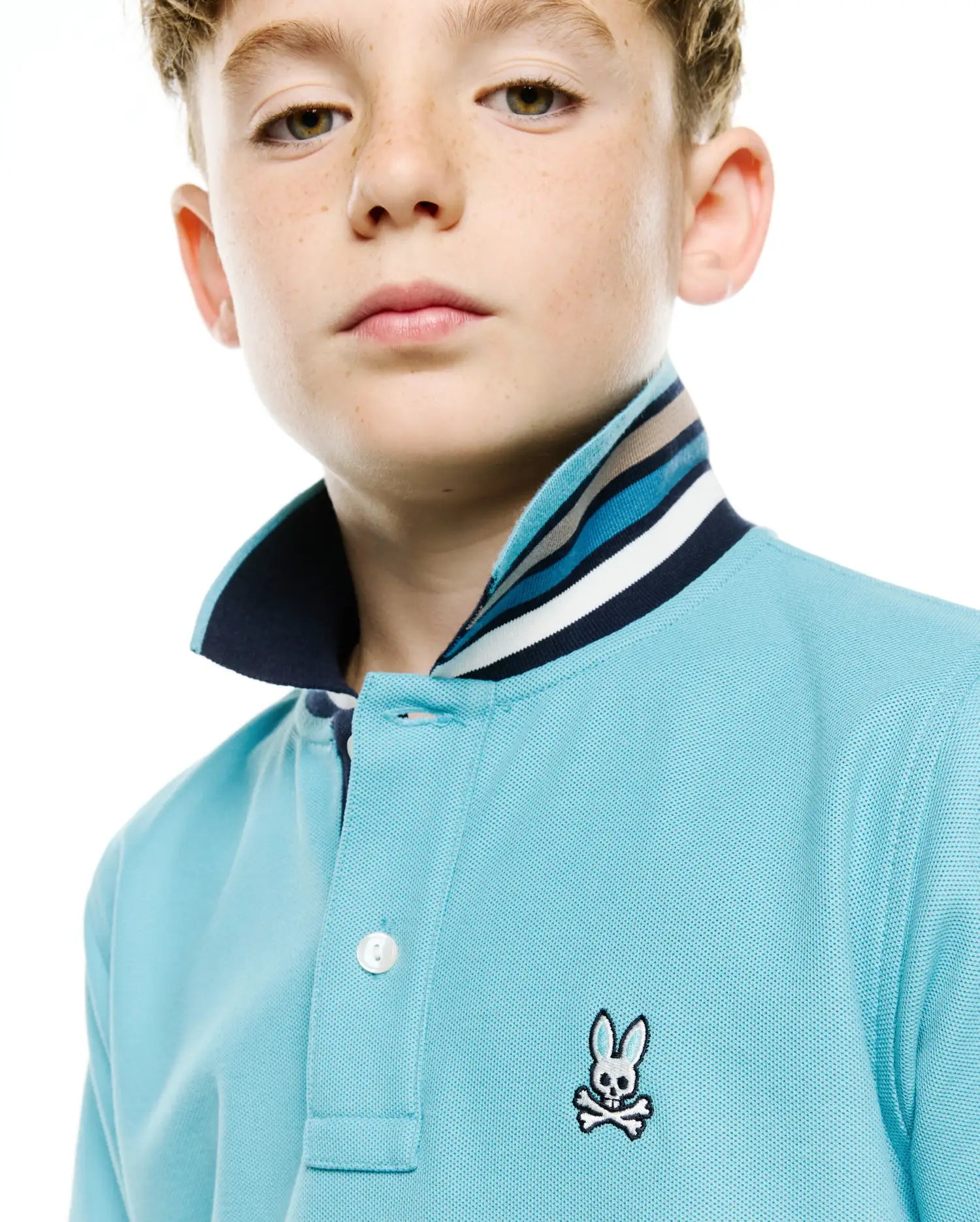 Kids' Klaus Pique Polo in light blue, featuring a classic fit, textured pique fabric, and a striped collar in navy, light blue, white, and beige. Small embroidered Psycho Bunny skull logo on the left chest. (2)