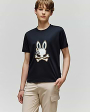 A young person stands against a plain background, wearing the Psycho Bunny KIDS WADE GRAPHIC TEE—an all-black, 100% cotton shirt with an embroidered Bunny head above crossed bones—and light beige pants featuring a thigh pocket.