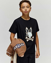 A young boy with short curly hair wears the Psycho Bunny KIDS WADE GRAPHIC TEE (KT0200054), featuring a large embroidered Bunny skull and crossbones. He holds a brown jacket over his left arm against a plain light background.