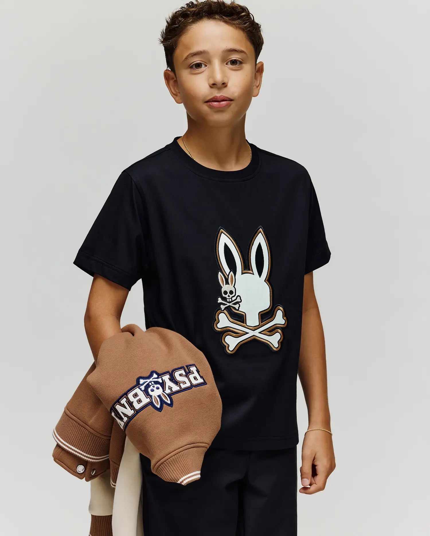 A young boy with short curly hair wears the Psycho Bunny KIDS WADE GRAPHIC TEE (KT0200054), featuring a large embroidered Bunny skull and crossbones. He holds a brown jacket over his left arm against a plain light background.