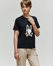 A young person with short brown hair wears the Psycho Bunny KIDS WADE GRAPHIC TEE - KT0200054, featuring a large embroidered Bunny with crossed bones, paired with beige cargo pants and standing against a plain light gray background.
