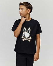 A boy with short brown hair wears the Psycho Bunny KIDS WADE GRAPHIC TEE (KT0200054), a black 100% cotton shirt with an embroidered Bunny skull and crossbones, as he looks to his right against a plain light background.