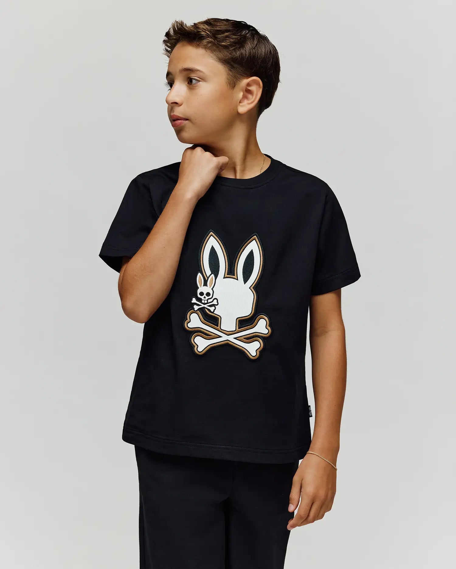 A boy with short brown hair wears the Psycho Bunny KIDS WADE GRAPHIC TEE (KT0200054), a black 100% cotton shirt with an embroidered Bunny skull and crossbones, as he looks to his right against a plain light background.