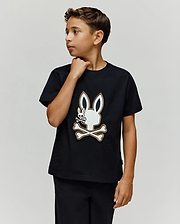 A boy with short brown hair wears the Psycho Bunny KIDS WADE GRAPHIC TEE (KT0200054), a black 100% cotton shirt with an embroidered Bunny skull and crossbones, as he looks to his right against a plain light background.