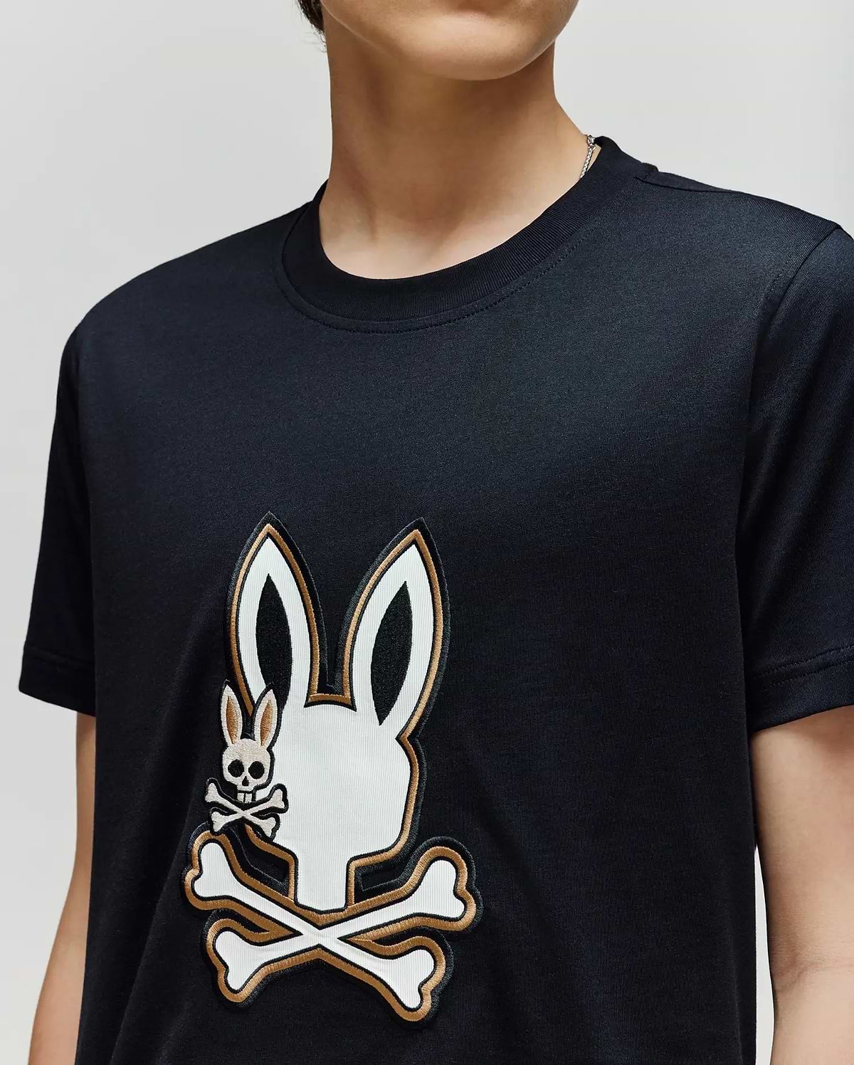 A person wearing the Psycho Bunny KIDS WADE GRAPHIC TEE (KT0200054), a black 100% cotton shirt with a large white bunny head and crossbones graphic and a smaller bunny skull and crossbones on the chest.