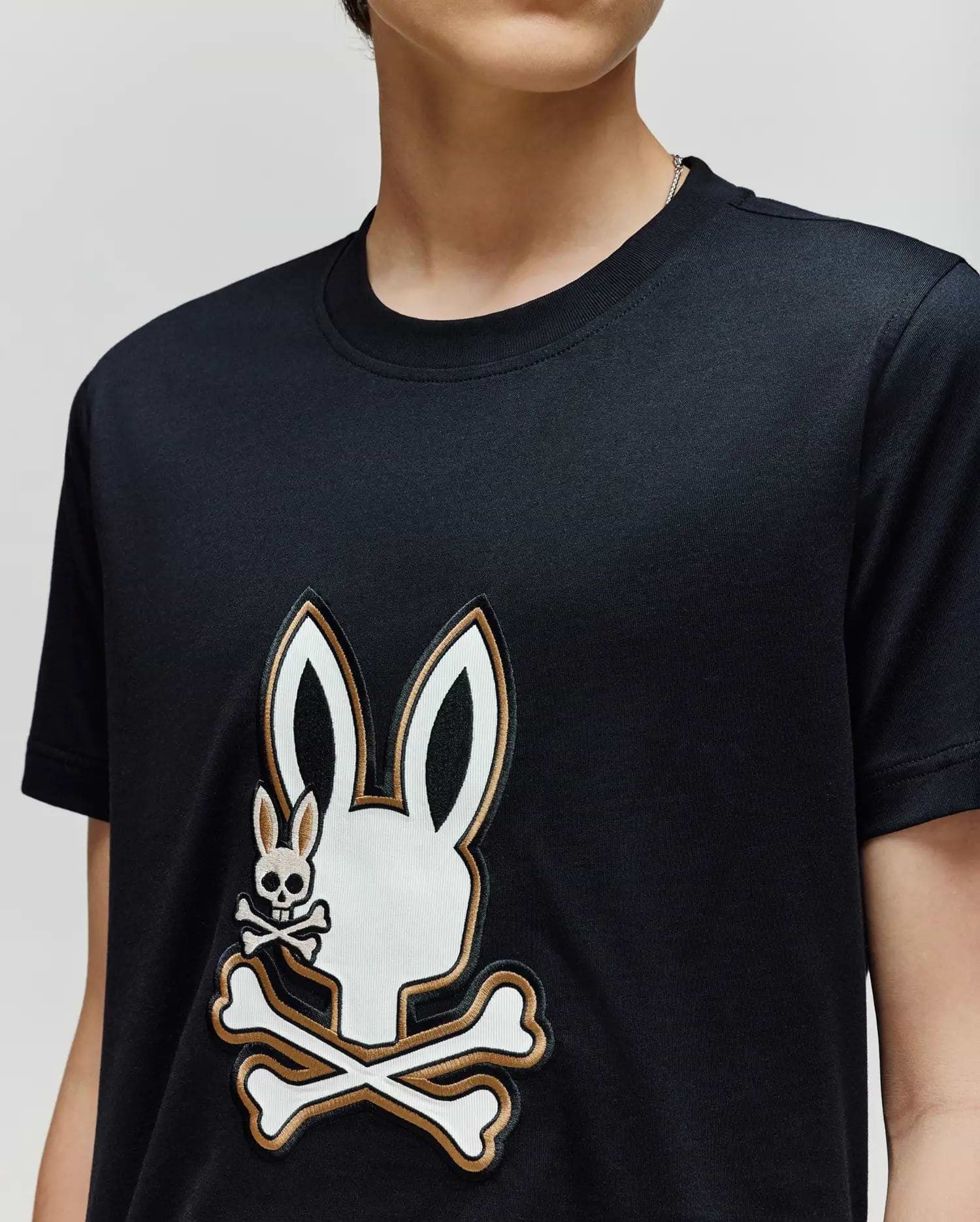 A person wearing the Psycho Bunny KIDS WADE GRAPHIC TEE (KT0200054), a black 100% cotton shirt with a large white bunny head and crossbones graphic and a smaller bunny skull and crossbones on the chest.