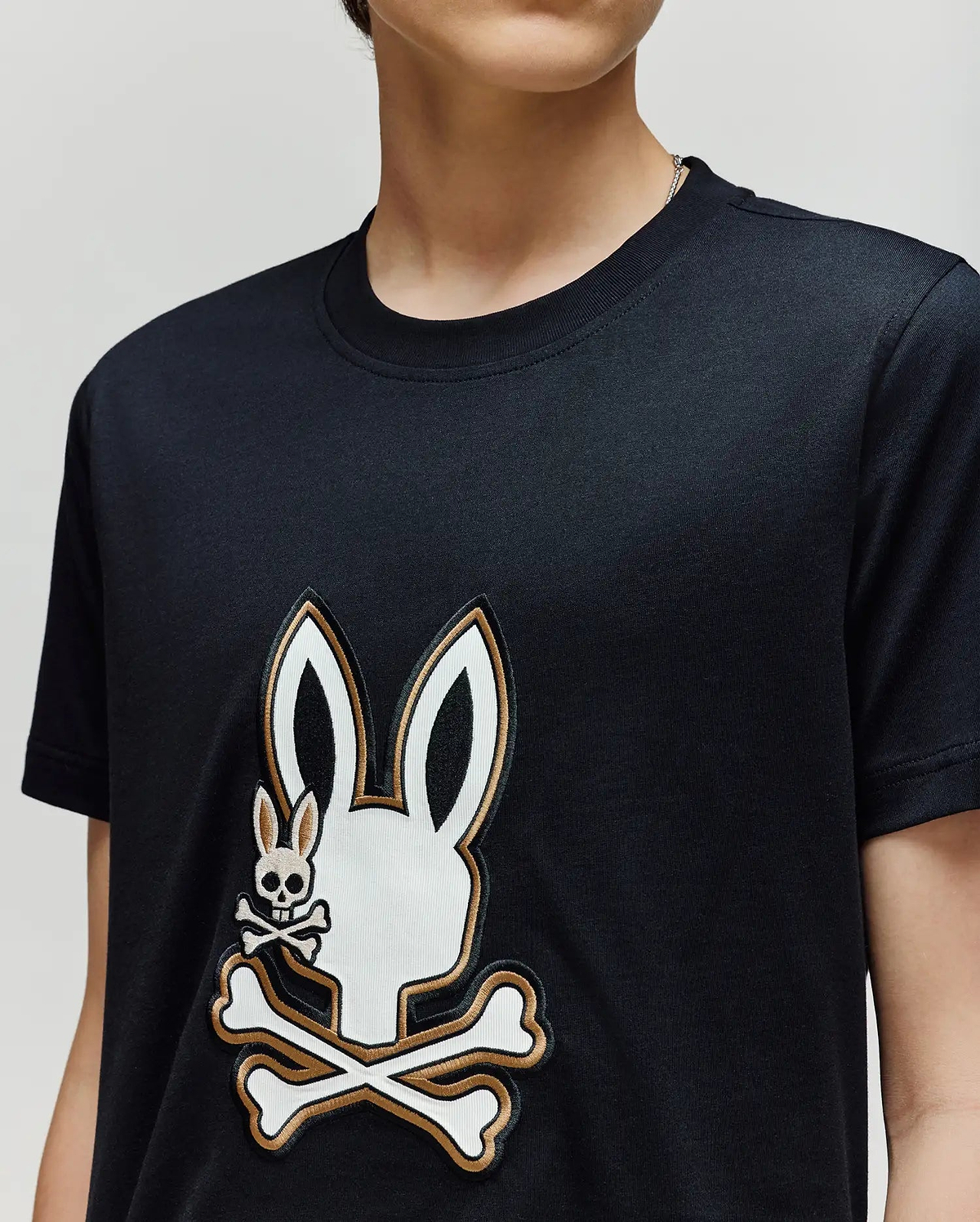 A person wearing the Psycho Bunny KIDS WADE GRAPHIC TEE (KT0200054), a black 100% cotton shirt with a large white bunny head and crossbones graphic and a smaller bunny skull and crossbones on the chest.