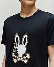 A person wearing the Psycho Bunny KIDS WADE GRAPHIC TEE (KT0200054), a black 100% cotton shirt with a large white bunny head and crossbones graphic and a smaller bunny skull and crossbones on the chest.