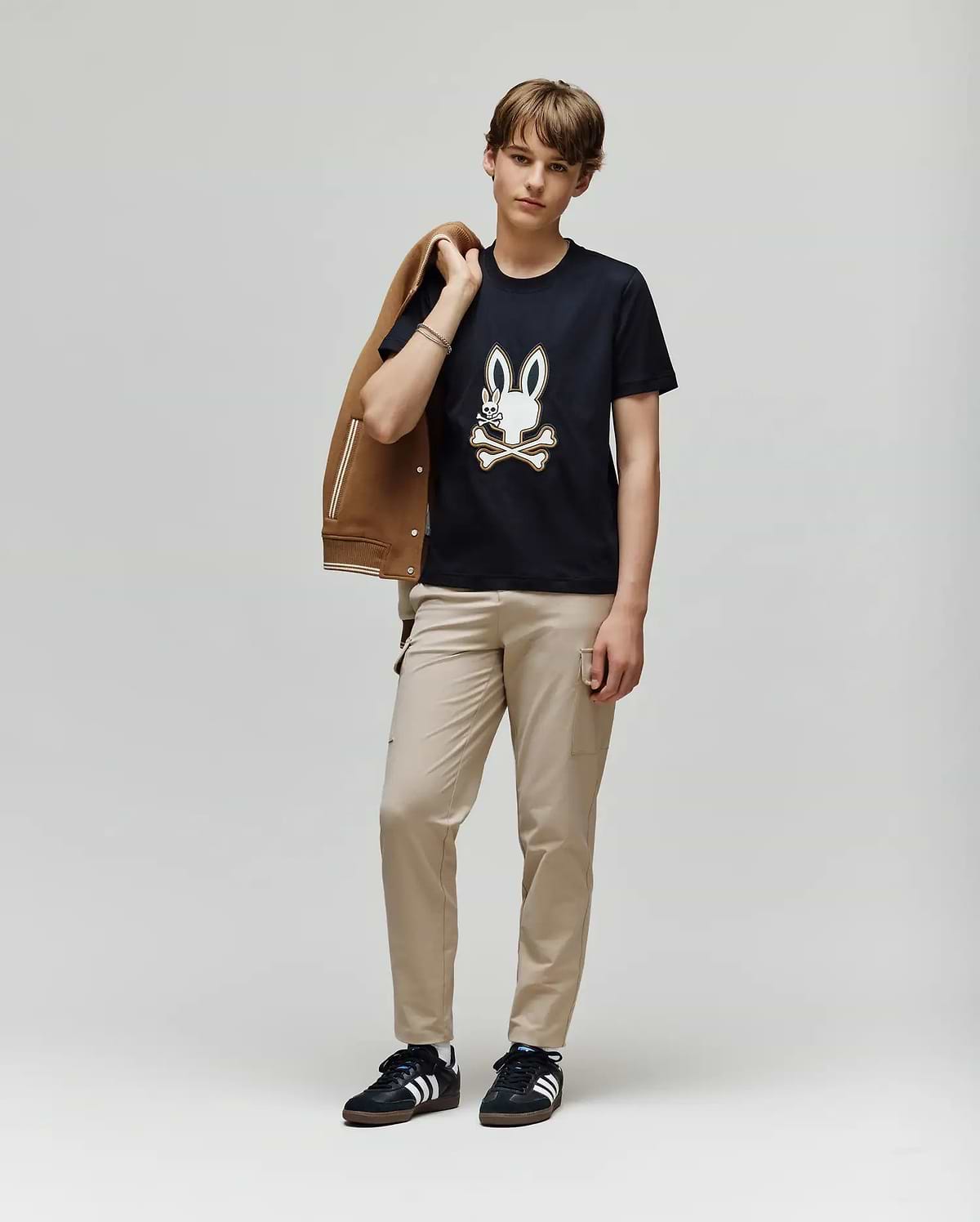 A young person stands against a plain light background, holding a brown jacket and wearing beige pants, black Adidas sneakers, and the Psycho Bunny KIDS WADE GRAPHIC TEE - KT0200054 with an embroidered Bunny and crossbones design.
