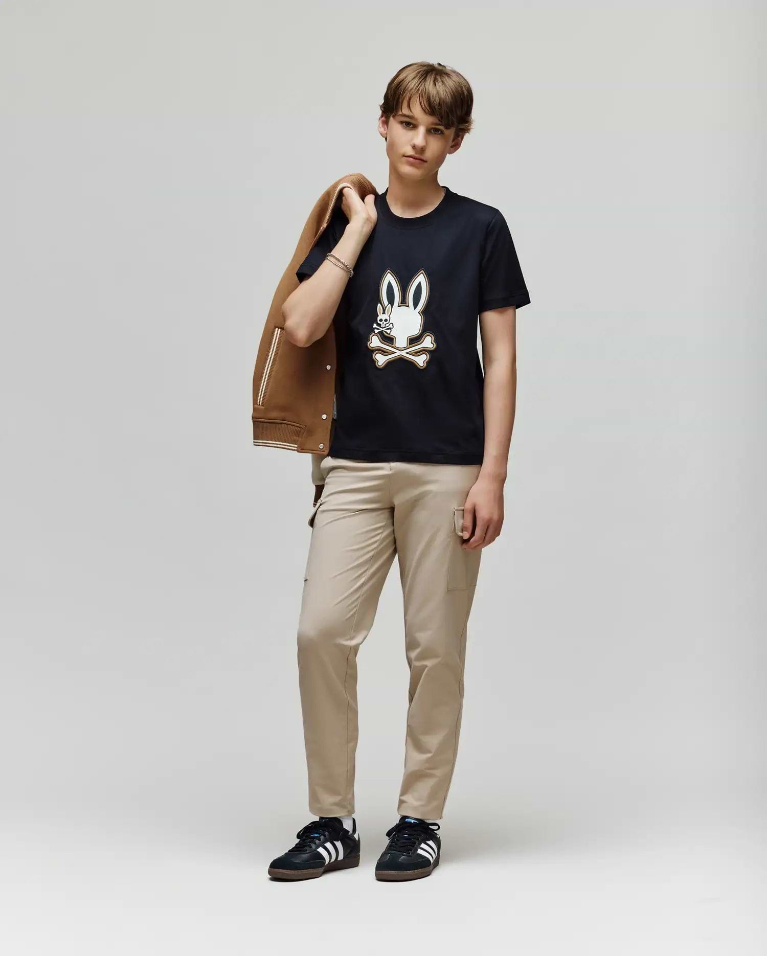 A young person stands against a plain light background, holding a brown jacket and wearing beige pants, black Adidas sneakers, and the Psycho Bunny KIDS WADE GRAPHIC TEE - KT0200054 with an embroidered Bunny and crossbones design.