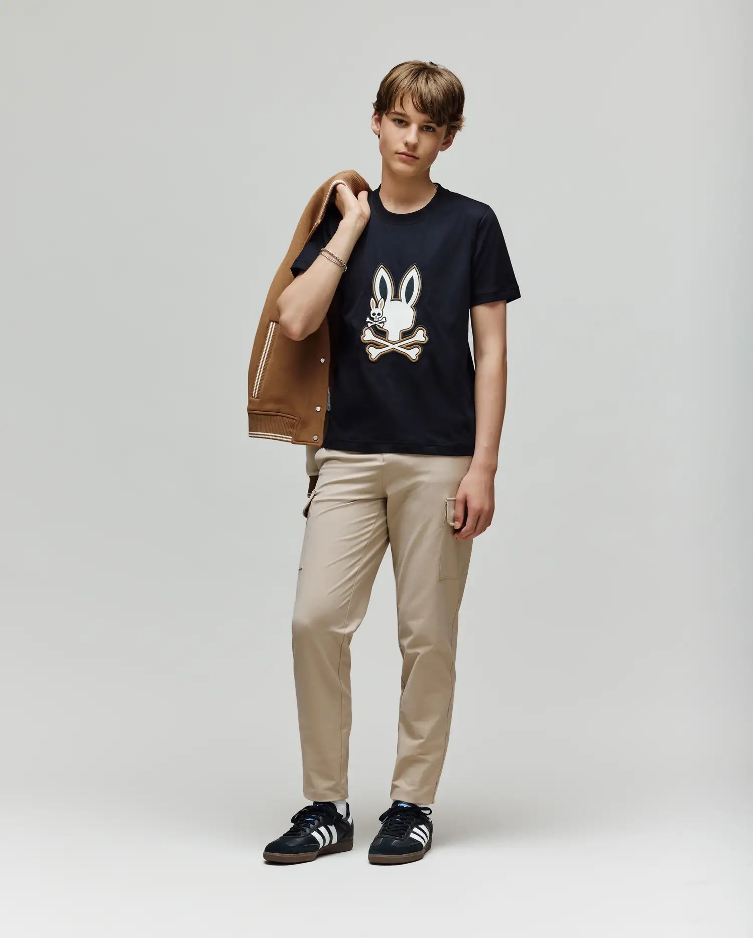A young person stands against a plain light background, holding a brown jacket and wearing beige pants, black Adidas sneakers, and the Psycho Bunny KIDS WADE GRAPHIC TEE - KT0200054 with an embroidered Bunny and crossbones design.