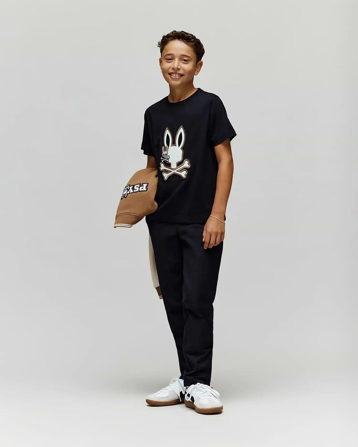A smiling boy stands against a plain background, wearing the Psycho Bunny KIDS WADE GRAPHIC TEE - KT0200054 with an embroidered bunny and crossbones, black pants, white sneakers, and holding a brown hat. The tee is made from 100% cotton for comfort.