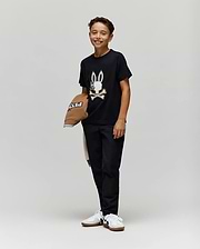 A smiling boy stands against a plain background, wearing the Psycho Bunny KIDS WADE GRAPHIC TEE - KT0200054 with an embroidered bunny and crossbones, black pants, white sneakers, and holding a brown hat. The tee is made from 100% cotton for comfort.