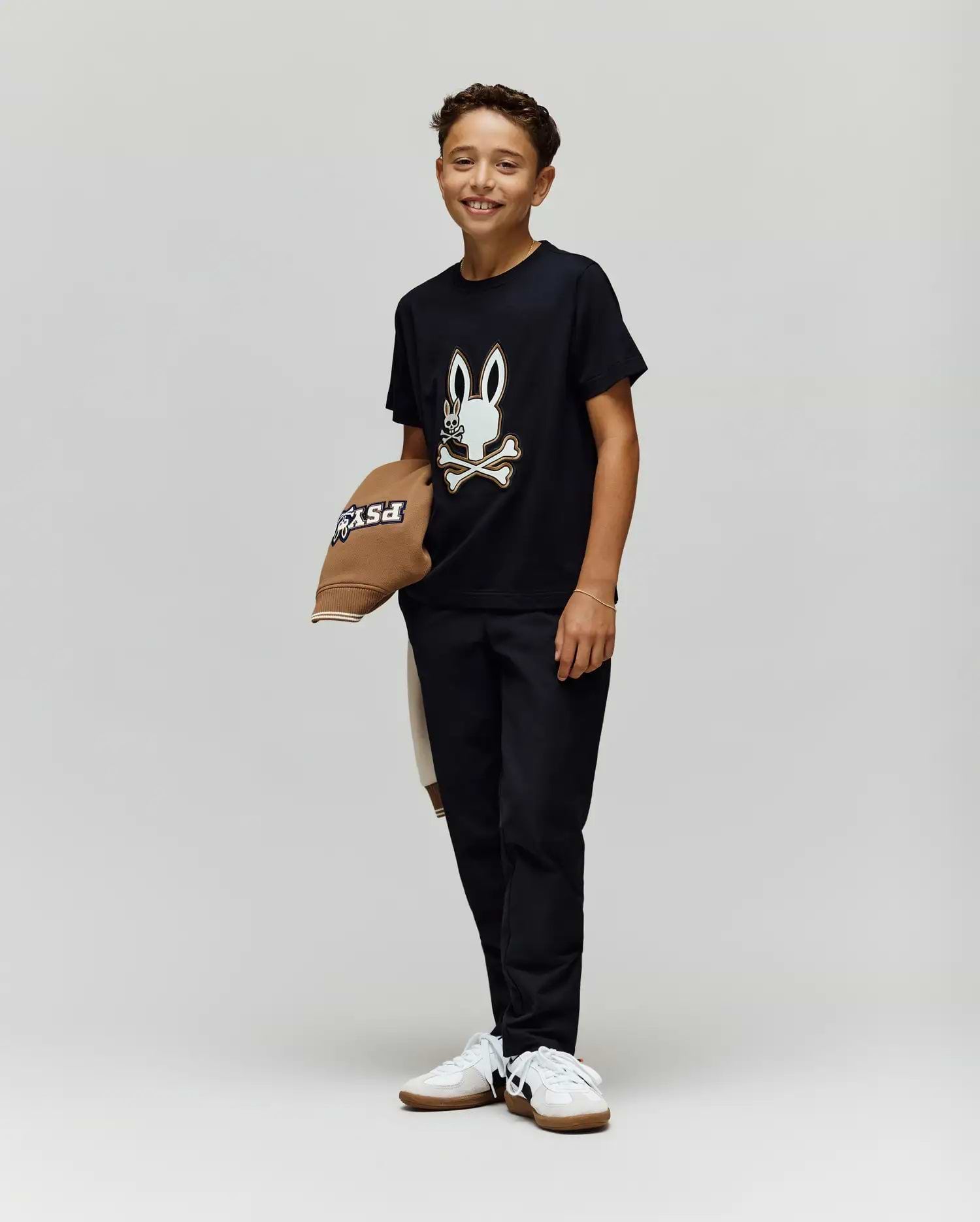 A smiling boy stands against a plain background, wearing the Psycho Bunny KIDS WADE GRAPHIC TEE - KT0200054 with an embroidered bunny and crossbones, black pants, white sneakers, and holding a brown hat. The tee is made from 100% cotton for comfort.