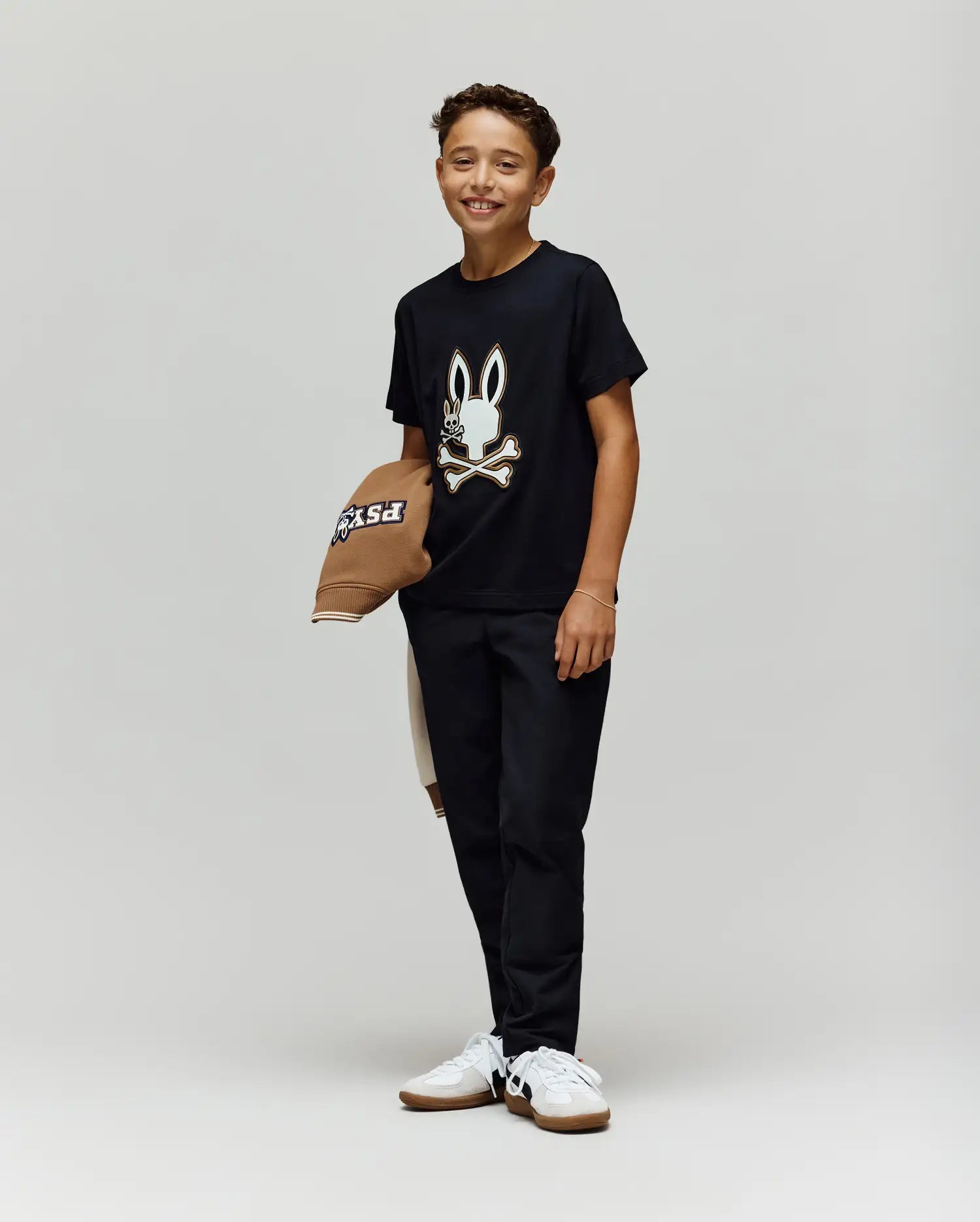 A smiling boy stands against a plain background, wearing the Psycho Bunny KIDS WADE GRAPHIC TEE - KT0200054 with an embroidered bunny and crossbones, black pants, white sneakers, and holding a brown hat. The tee is made from 100% cotton for comfort.