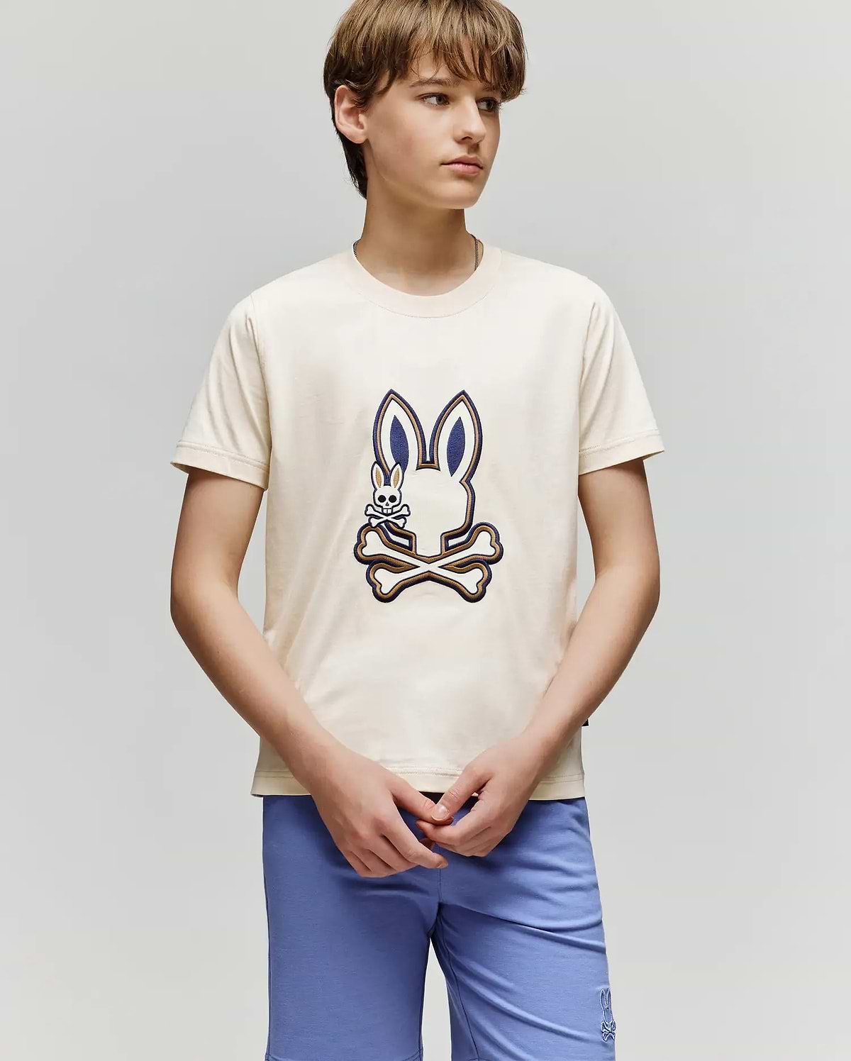 A teenage boy wears the Psycho Bunny KIDS WADE GRAPHIC TEE - KT0200054, a cream-colored shirt with an embroidered Bunny and crossbones design, paired with light blue shorts, standing against a plain light gray background.