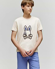 A teenage boy wears the Psycho Bunny KIDS WADE GRAPHIC TEE - KT0200054, a cream-colored shirt with an embroidered Bunny and crossbones design, paired with light blue shorts, standing against a plain light gray background.