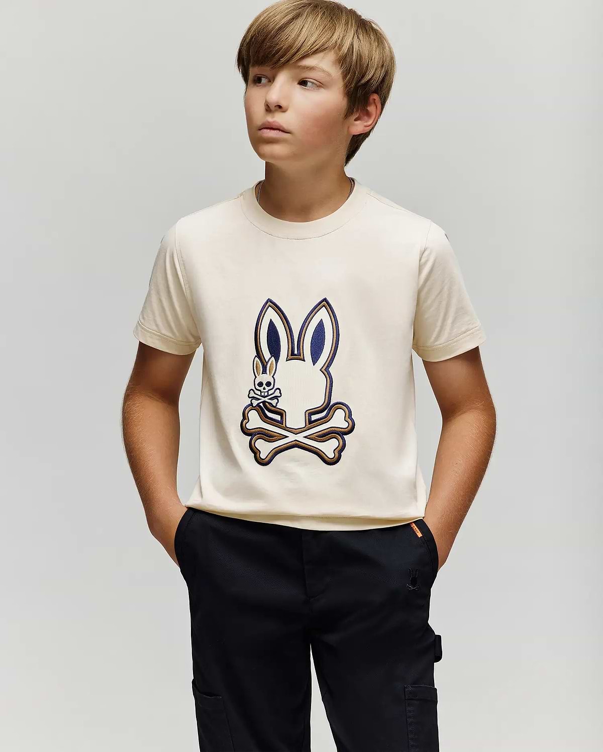 A boy with light brown hair wears the cream KIDS WADE GRAPHIC TEE - KT0200054 by Psycho Bunny, featuring an embroidered Bunny and crossbones. Made from 100% cotton, he pairs it with black pants and stands against a plain background.