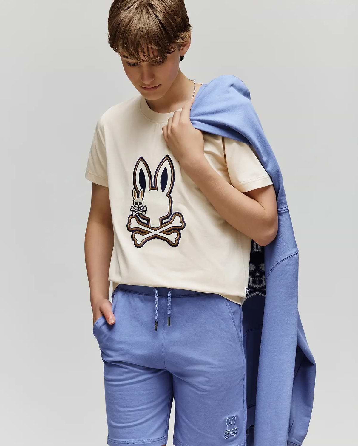 A child wears the KIDS WADE GRAPHIC TEE - KT0200054 by Psycho Bunny, a beige 100% cotton tee with an embroidered Bunny and crossbones, paired with blue shorts and a matching blue sweatshirt draped over one shoulder.