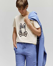 A child wears the KIDS WADE GRAPHIC TEE - KT0200054 by Psycho Bunny, a beige 100% cotton tee with an embroidered Bunny and crossbones, paired with blue shorts and a matching blue sweatshirt draped over one shoulder.