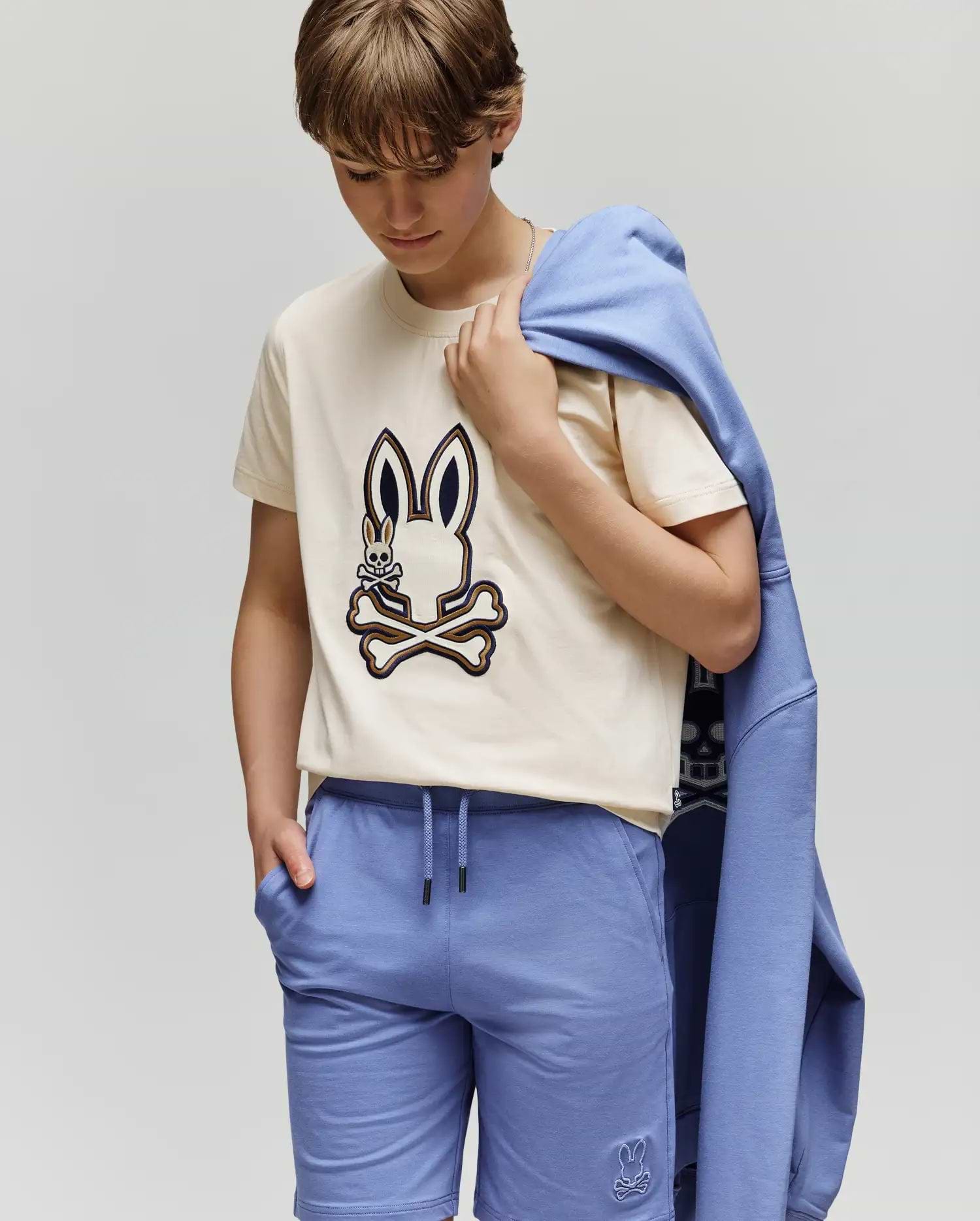 A child wears the KIDS WADE GRAPHIC TEE - KT0200054 by Psycho Bunny, a beige 100% cotton tee with an embroidered Bunny and crossbones, paired with blue shorts and a matching blue sweatshirt draped over one shoulder.