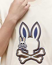 Someone wearing the Psycho Bunny KIDS WADE GRAPHIC TEE—KT0200054, a cream-colored 100% cotton shirt with a large embroidered Bunny head above crossed bones and a smaller bunny skull and crossbones on the left side.